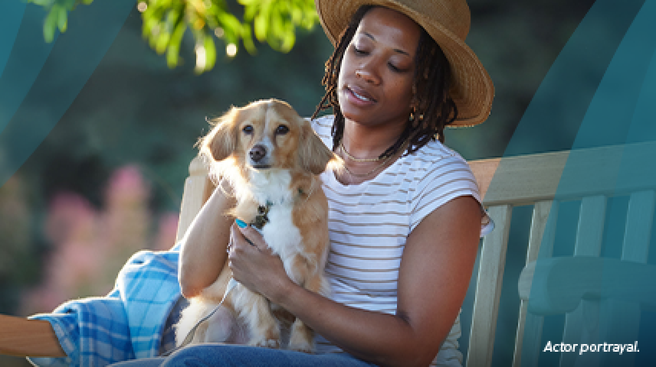 A Black woman in her 40s is sitting on a park bench outside, petting her small tan dog on her lap. The woman has chin‐length black curls and is wearing a sun hat. Actor portrayal.
