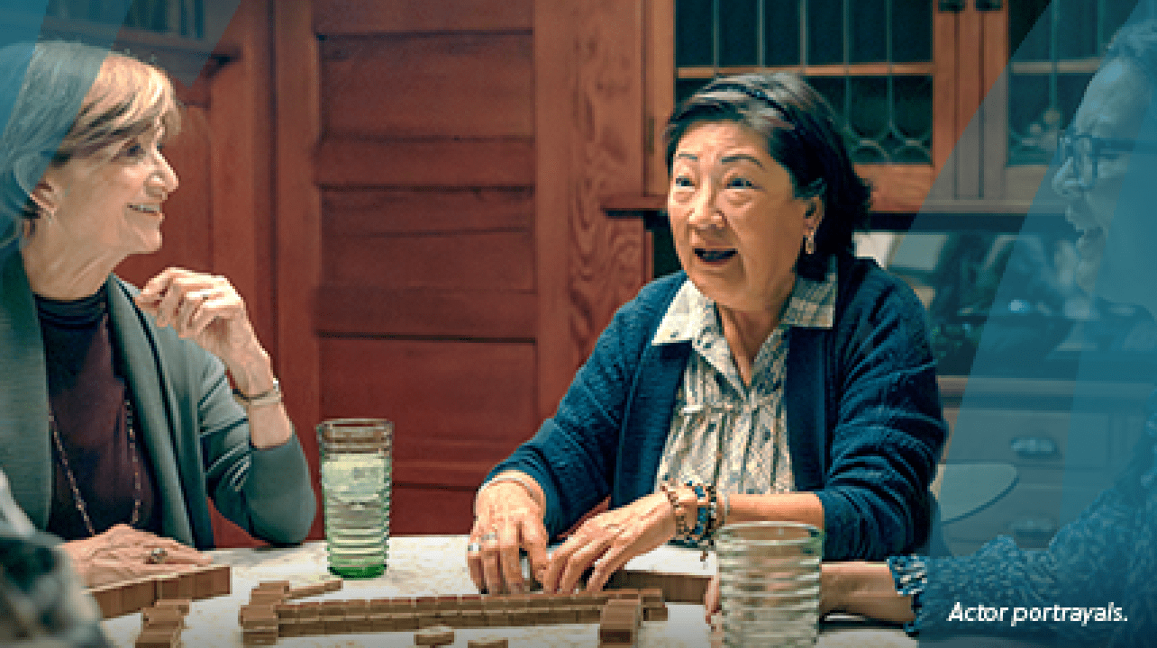 An Asian woman in her mid-60s with short dark hair is playing mahjong with a group of friends around a dining room table. They are all talking and smiling. Actor portrayals.