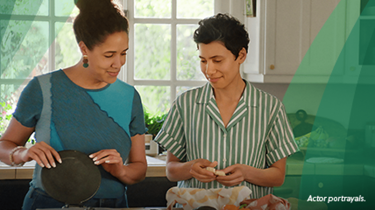 A Latina woman in her 40s with short, dark, curly hair stands in the kitchen with her partner, another Latina woman in her 40s. They are making tortillas from scratch, with one woman forming the masa and the other pressing it in the tortilla press. Actor portrayals.