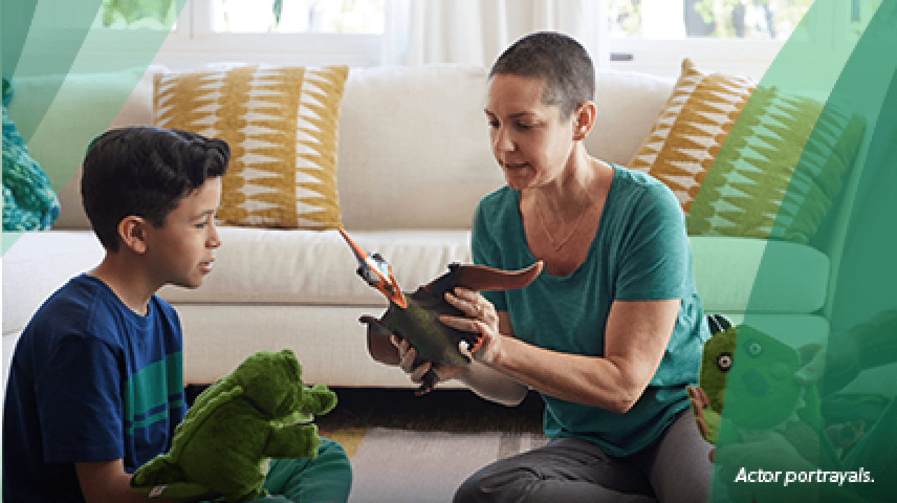 A White woman in her 30s with shaved dark hair sits on the living room floor in front of the couch between a White man in his 30s and a 7‐year‐old boy. The woman is holding a dinosaur action figure and is playing with the boy, who has a dinosaur puppet on his hand. Actor portrayals.