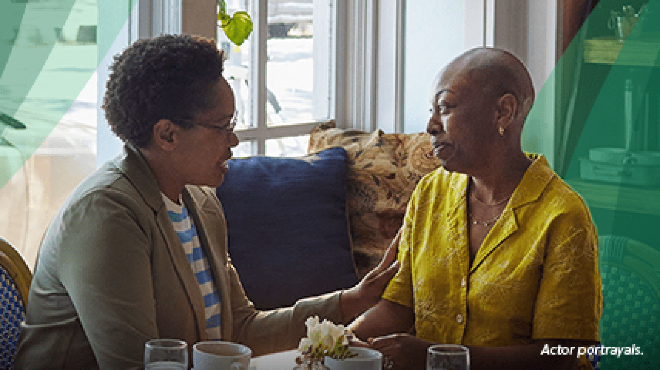 A Black woman in her 40s with closely shaved hair sits at a table in a coffee shop with her friend, another Black woman in her 40s. The friend is gently holding the other woman's arm in a gesture of support. Actor portrayals.