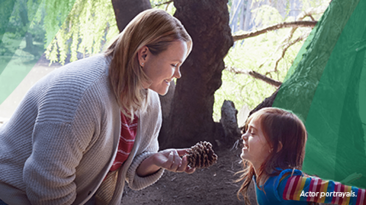 A White woman in her 30s with shoulder‐length, blonde, straight hair is out on a nature walk with her 6‐year‐old daughter. The woman is smiling and holding a pine cone that her daughter found, as the girl smiles and jumps excitedly. Actor portrayals