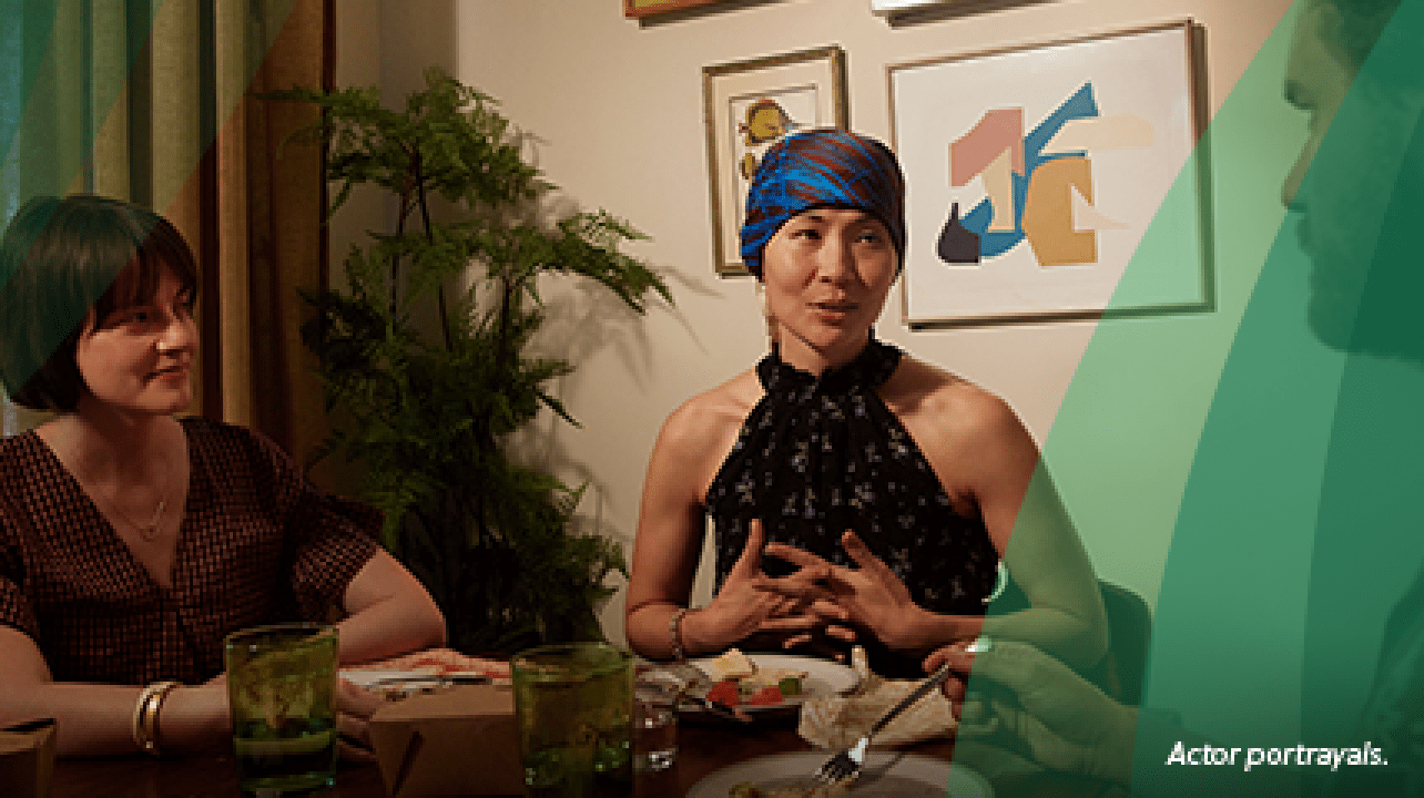 An Asian woman in her 40s wearing a red and blue silk headscarf sits at a round dining table, smiling and talking with 2 friends, a White man and White woman of a similar age. They are finishing dinner, a takeout meal from one of their favorite restaurants. Actor portrayals.