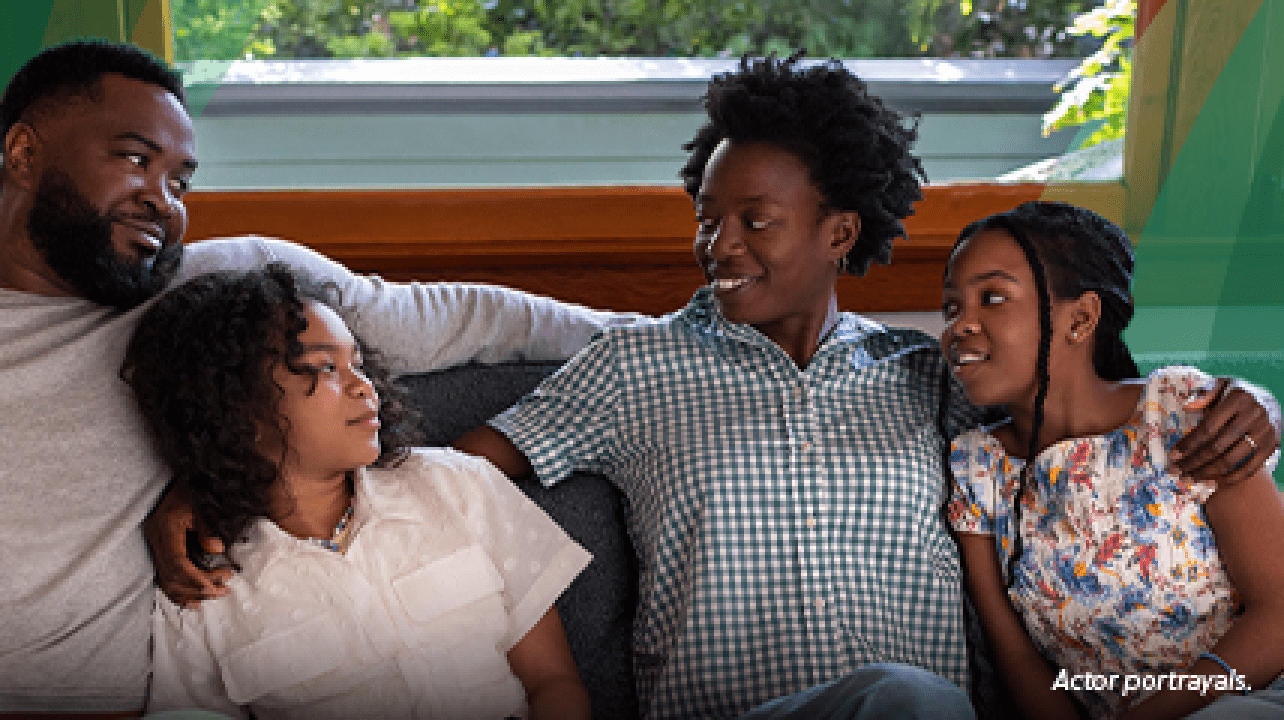 A Black woman in her 40s with short natural hair is cuddled on the living room couch with her family‐‐ a Black man in his 40s and their 2 preteen daughters. The woman has her arms around both of her daughters and is smiling at her husband. The family is close, leaning on each other and smiling. Actor portrayals.
