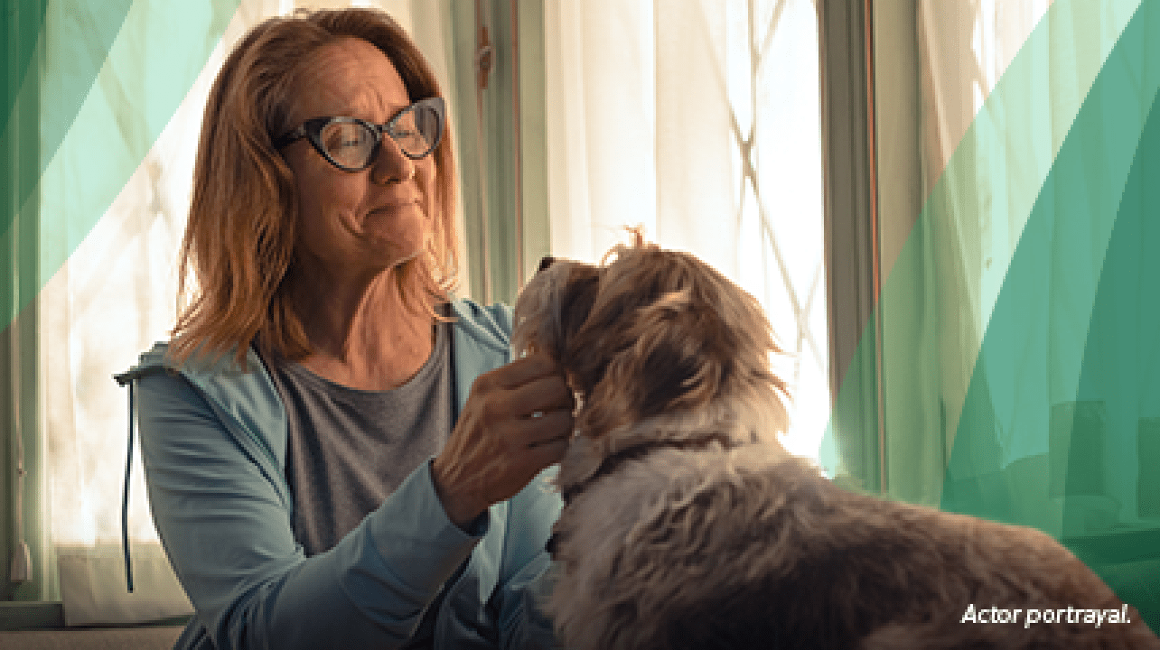 A White woman in her 50s with shoulder‐length blonde hair and glasses is sitting on her couch and smiling softly as she pets a medium‐sized tan and White dog with shaggy fur. Actor portrayal.